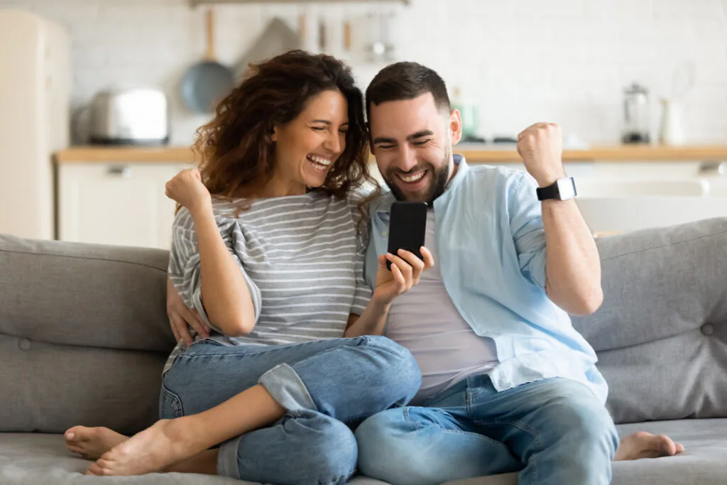 A couple on their couch at home celebrating a winning bid on a phone