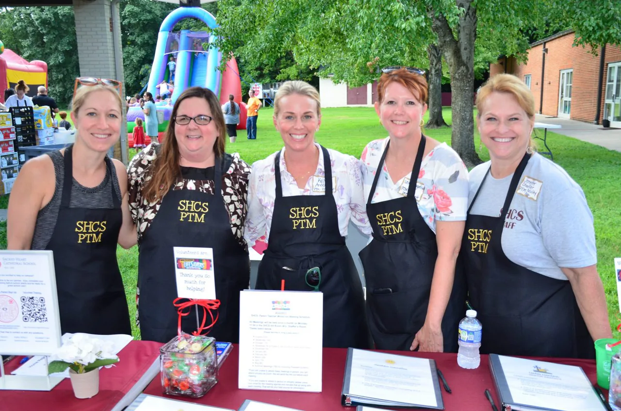 School parent-teacher volunteers smiling behind their table at an outdoor school fundraiser