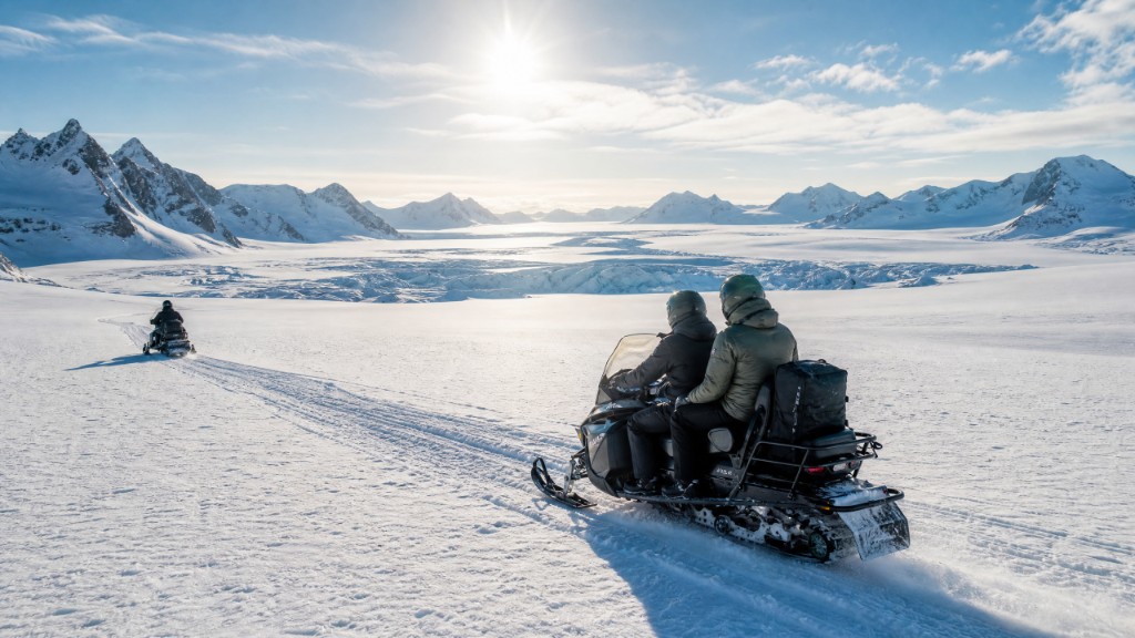 Snowmobilers crossing a sunlit glacier with mountain peaks ahead